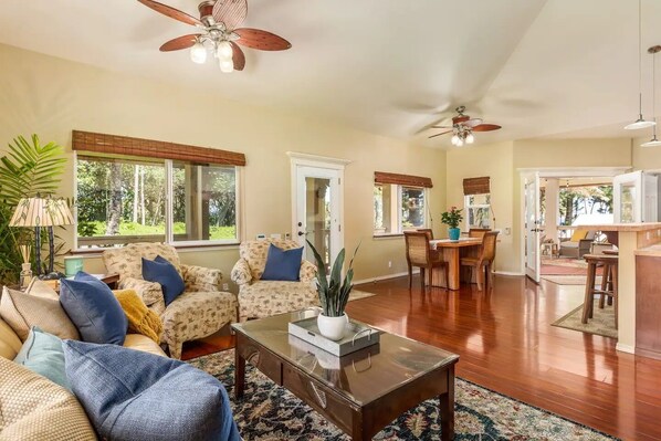 A spacious living room with a wooden coffee table, two patterned armchairs, a dining area with a wooden table, and a kitchen area visible through an open door. - Moana Imaku - Ocean View Home w/ ADA Features (Pahoa)
