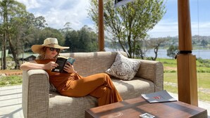 A person sitting on a beige sofa with a patterned pillow, reading a book. A laptop is on a wooden table beside the sofa. - W15 Lake Gergory (Nuwara Eliya)