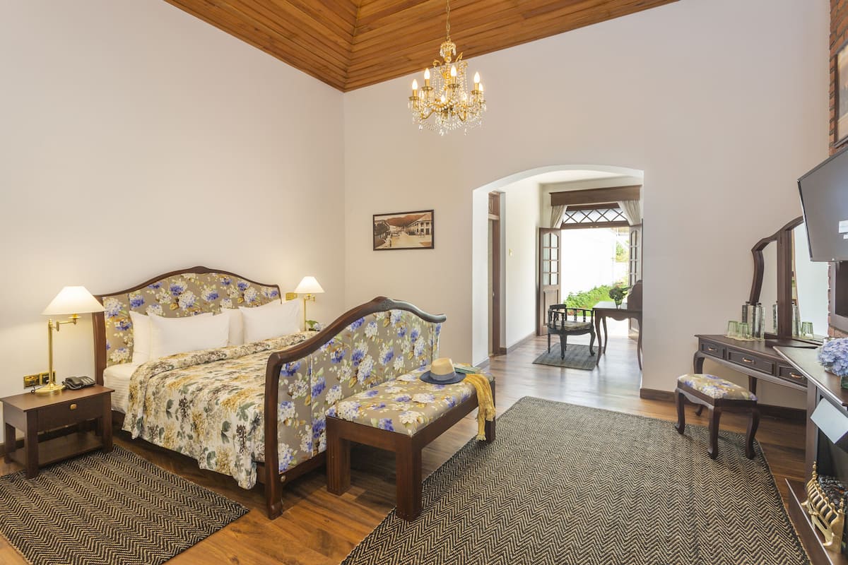 Un dormitorio con una cama de madera, dos mesitas de noche con lámparas, un banco, un escritorio con silla y una araña.