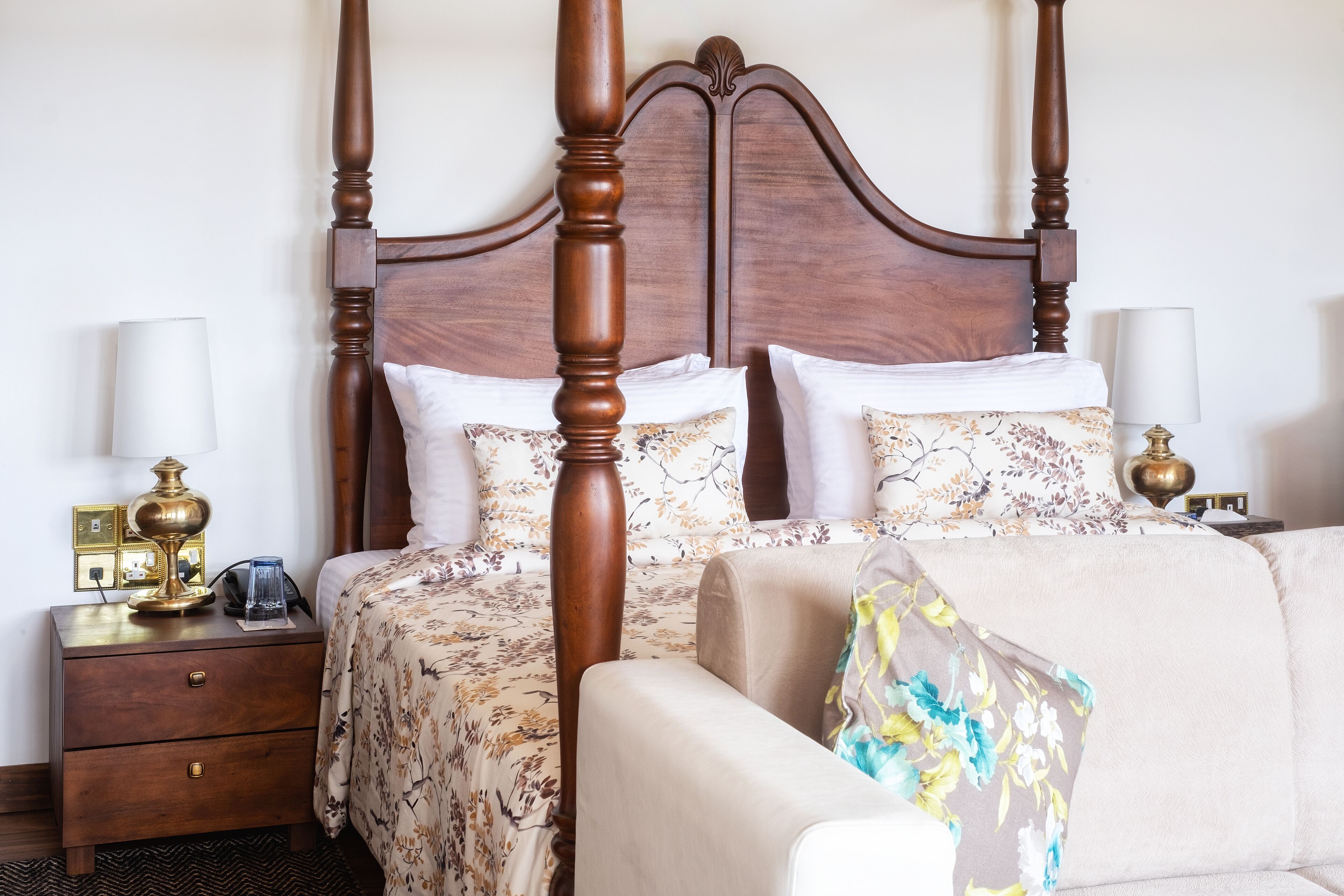 Une chambre d’hôtel avec un lit à baldaquin, une table de chevet avec une lampe et une chaise agrémentée d’un coussin à motifs floraux.