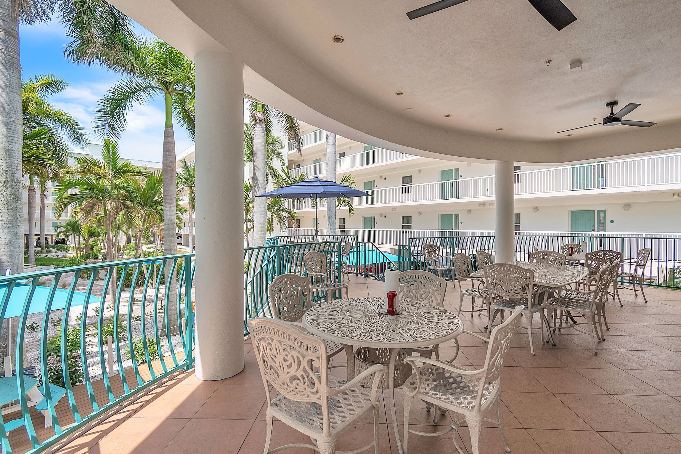 A covered outdoor area with tables and chairs, a pool, and a view of palm trees and a building.