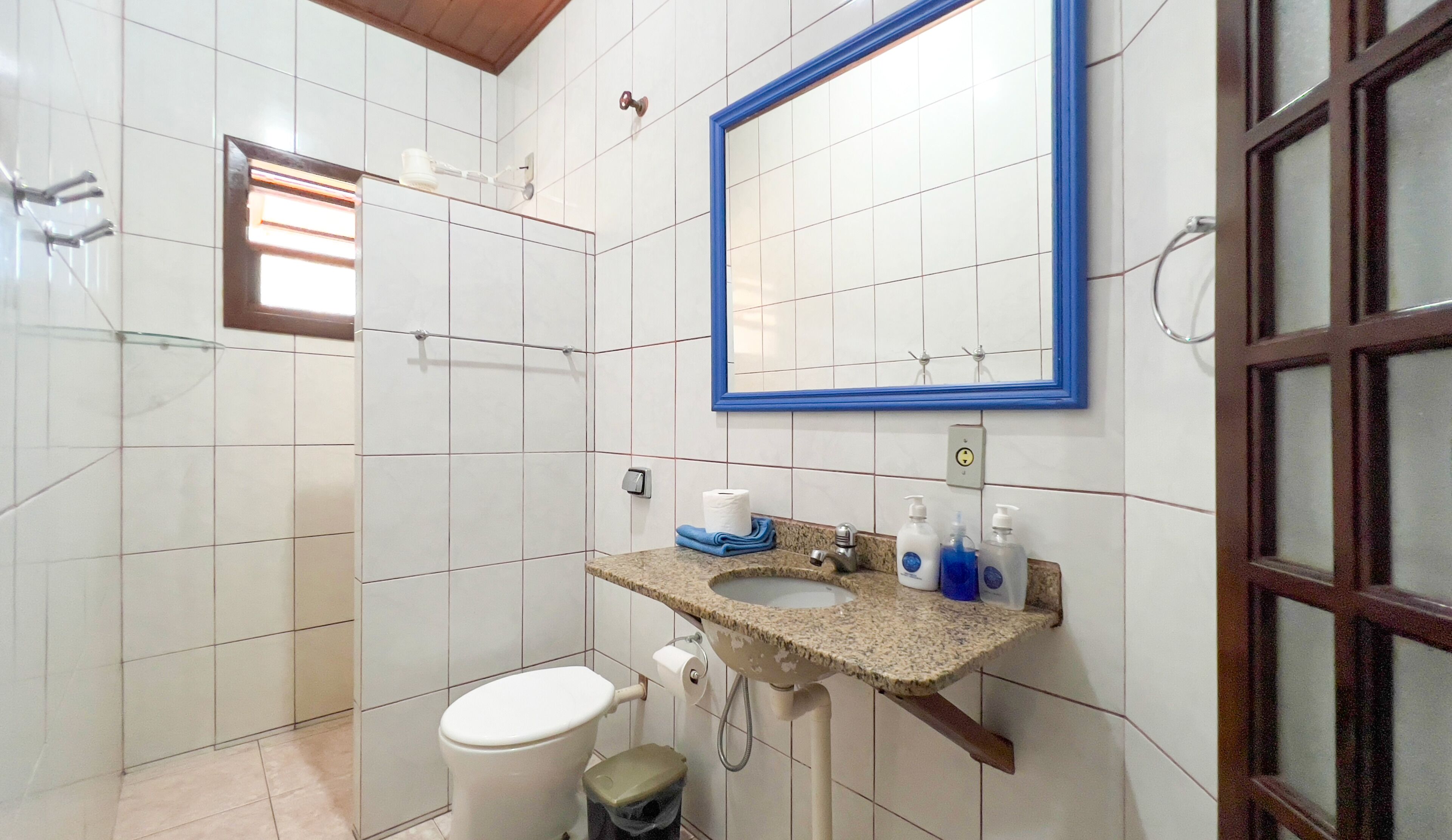 Un baño con paredes de azulejos blancos, un lavamanos con dos grifos, un espejo con marco azul, un inodoro y un cubo de basura.