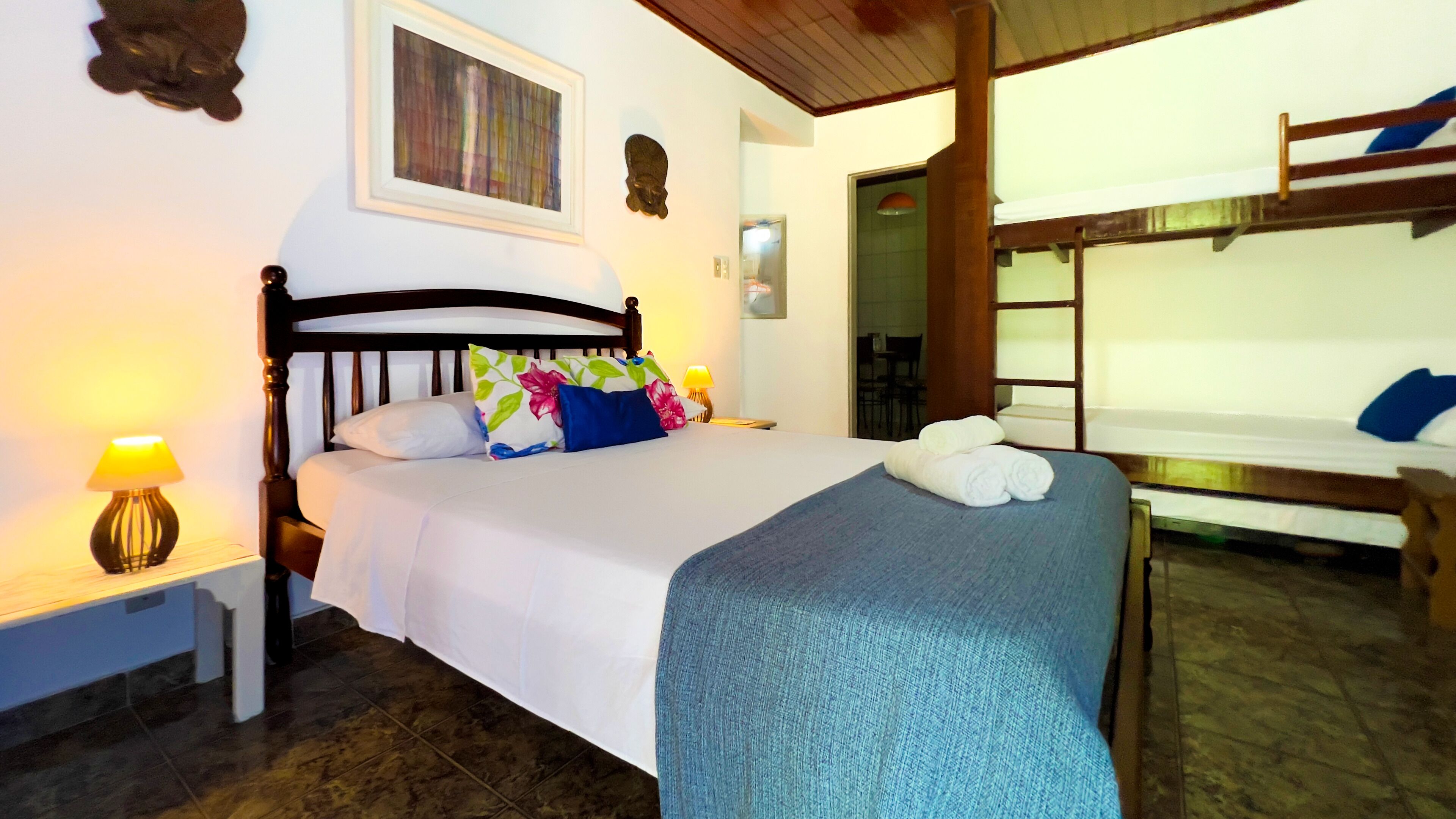 A hotel room with a bed, a nightstand, a lamp, a painting, and a bunk bed.
