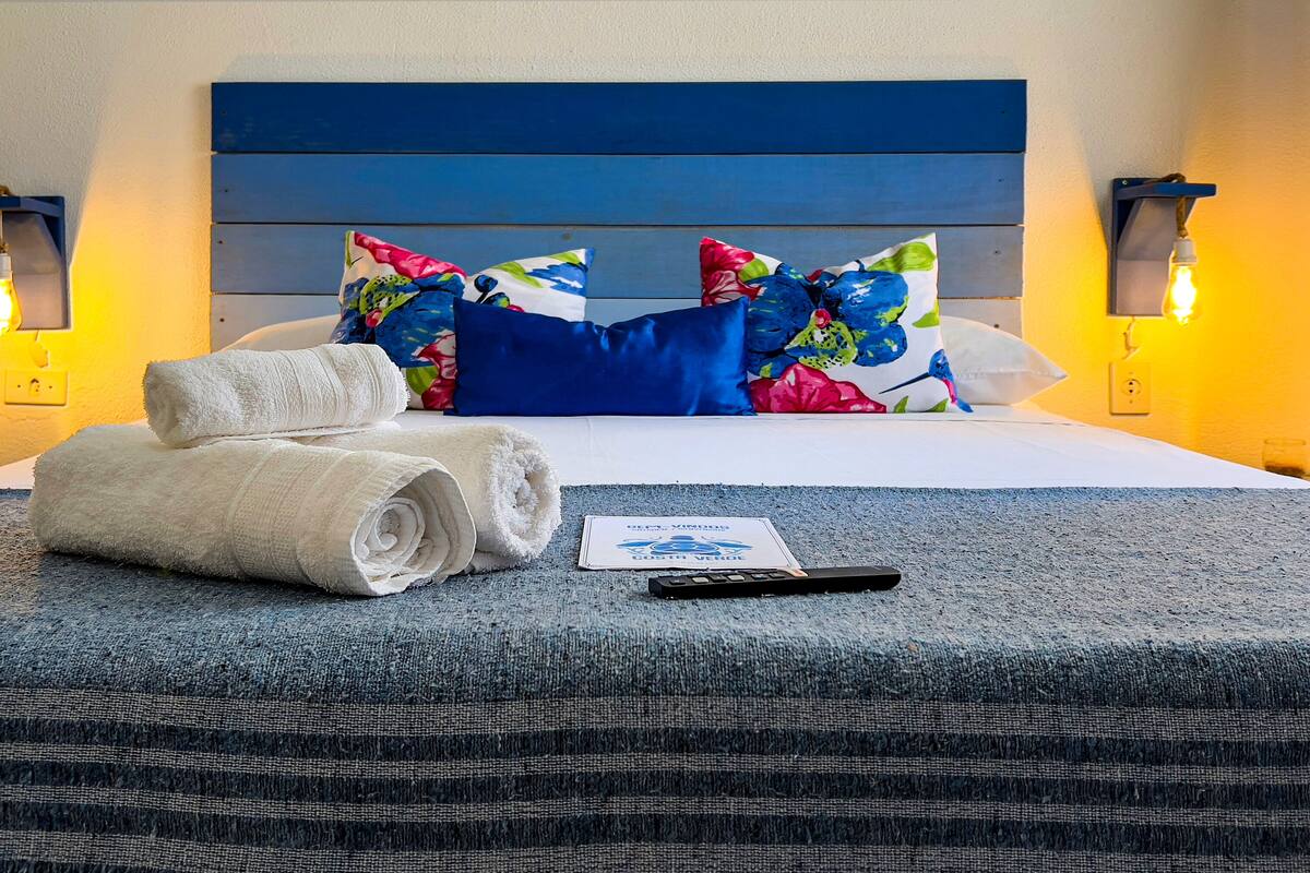 A neatly made bed with blue and floral pillows, two rolled towels, a remote control, and a card on a blue textured bedspread.