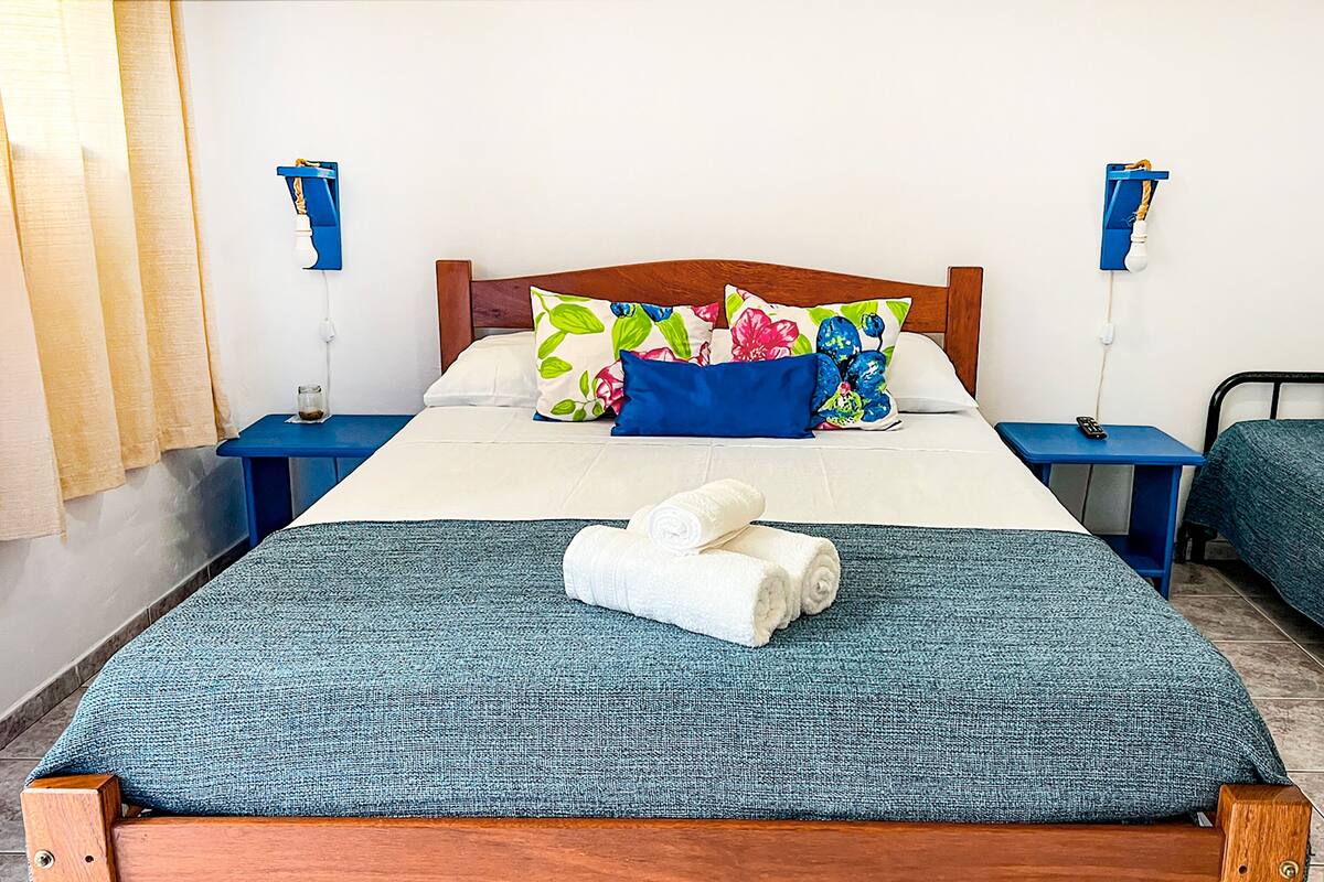 A double bed with a blue bedspread, two pillows, a blue nightstand, a bedside table with a lamp, and a window with curtains.