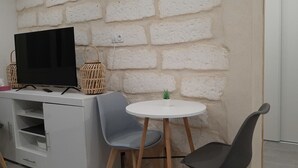 Ein kleiner, moderner Raum mit weißer Ziegelwand, einem Flachbildfernseher auf einem weißen Schrank, zwei grauen Stühlen um einen kleinen weißen Tisch und einer kleinen Topfpflanze auf dem Tisch.