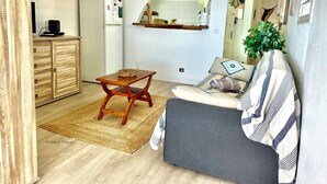 A living room with a sofa, a wooden coffee table, a rug, a clock, a refrigerator, and a small kitchen area with a shelf and a hanging sign. - Ti Cabanon. Sea view apartment in the center of Deshaies (Deshaies)
