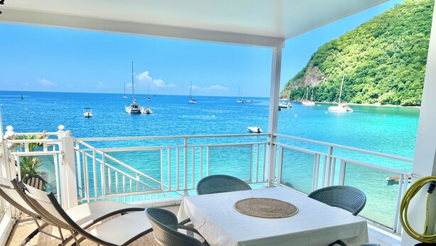 A balcony with a table and chairs, overlooking a turquoise sea with boats. - Ti Cabanon. Sea view apartment in the center of Deshaies (Deshaies)