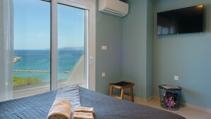 A bedroom with a bed, a small stool, a side table with magazines, and a TV on the wall. - Indigo Luxury apartment (Agios Nikolaos)