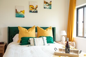 A neatly made bed with green and yellow pillows, a bedside table with a lamp and wine glasses, and artwork on the wall. - Luxe High Rise Uptown Condo |Free Wi-Fi | Secure Building | Free Parking (Charlotte)