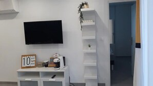 A room with a flat-screen TV mounted on the wall, a small desk with a coffee maker, a sign reading "10 AOU", and a plant on a shelf. - Happy Home for You Lunel: Happiness at the end of the journey. (Lunel)