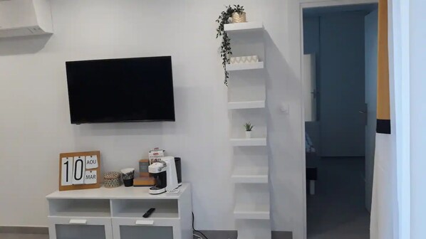 A room with a flat-screen TV mounted on the wall, a small desk with a coffee maker, a sign reading "10 AOU", and a plant on a shelf.