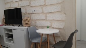 A small, modern room with a white brick wall, a flat-screen TV on a white cabinet, two grey chairs around a small white table, and a small potted plant on the table.