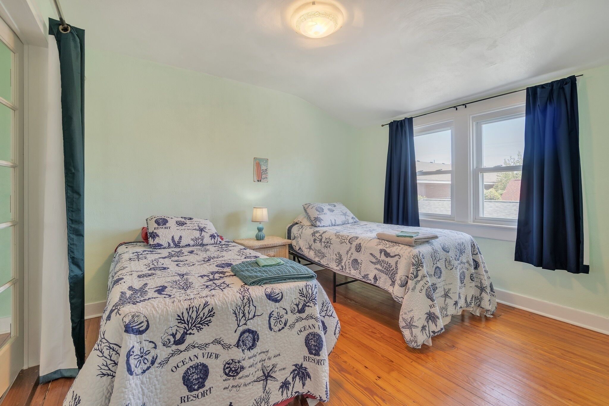 Habitación con dos camas, cada una con una colcha azul y blanca de temática marina. Cuenta con una ventana con cortinas azules, una mesita con una lámpara y un cuadro colgado en la pared.