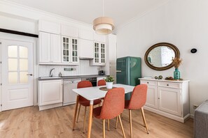 Une cuisine moderne avec des meubles blancs, un réfrigérateur vert de style rétro et un coin repas comprenant une table blanche et des chaises orange.
