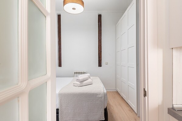 Un couloir étroit mène à une chambre qui contient un lit, une armoire et un luminaire suspendu.