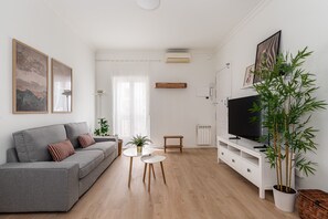 A modern living room with a grey sofa, a small round table, a TV on a white stand, and a large bamboo plant. - Waou Malasaña IV - Alquiler Corta Duración (Madrid)