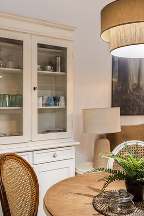 A dining area with a round wooden table, a wicker chair, a cabinet with glass doors, and a hanging lamp.