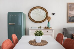 Un coin repas avec une table blanche, des chaises rouges, un réfrigérateur de couleur turquoise, un meuble blanc agrémenté d’objets décoratifs et un miroir rond accroché au mur.