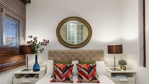 Een slaapkamer met een geborduurde hoofdbord, twee nachtkastjes, een ronde spiegel, een lamp, een vaas met bloemen en een bed met gekleurde kussens.