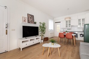 A modern living room with a flat-screen TV, a dining area with red chairs, and a kitchen with white cabinets.
