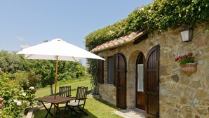 A stone building with arched windows and a door, a wooden table with chairs, and a white umbrella. - Cozy retreat for 2 with pool & A/C in Val d'Orcia | Moraiolo  (Monticchiello)