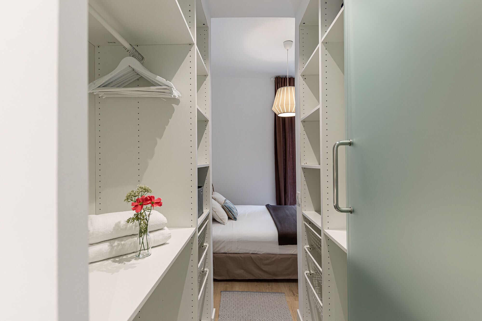 Un armario estrecho empotrado con estantes metálicos, una cama con almohadas, un florero con flores y una lámpara colgante.