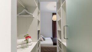 Un petit dressing étroit avec des étagères en métal, un lit avec des oreillers, un vase avec des fleurs et un luminaire suspendu.