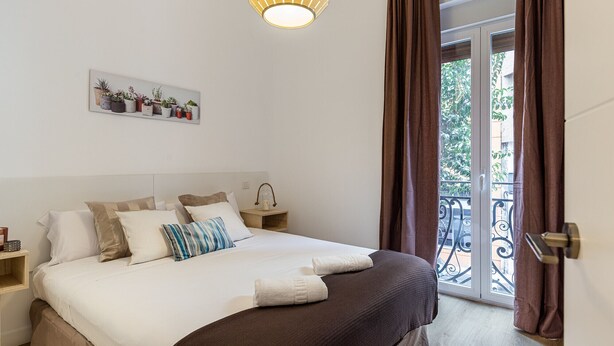 Une chambre à coucher avec un lit, des oreillers, une table de chevet, une lampe, une fenêtre avec des rideaux et une vue sur le balcon.