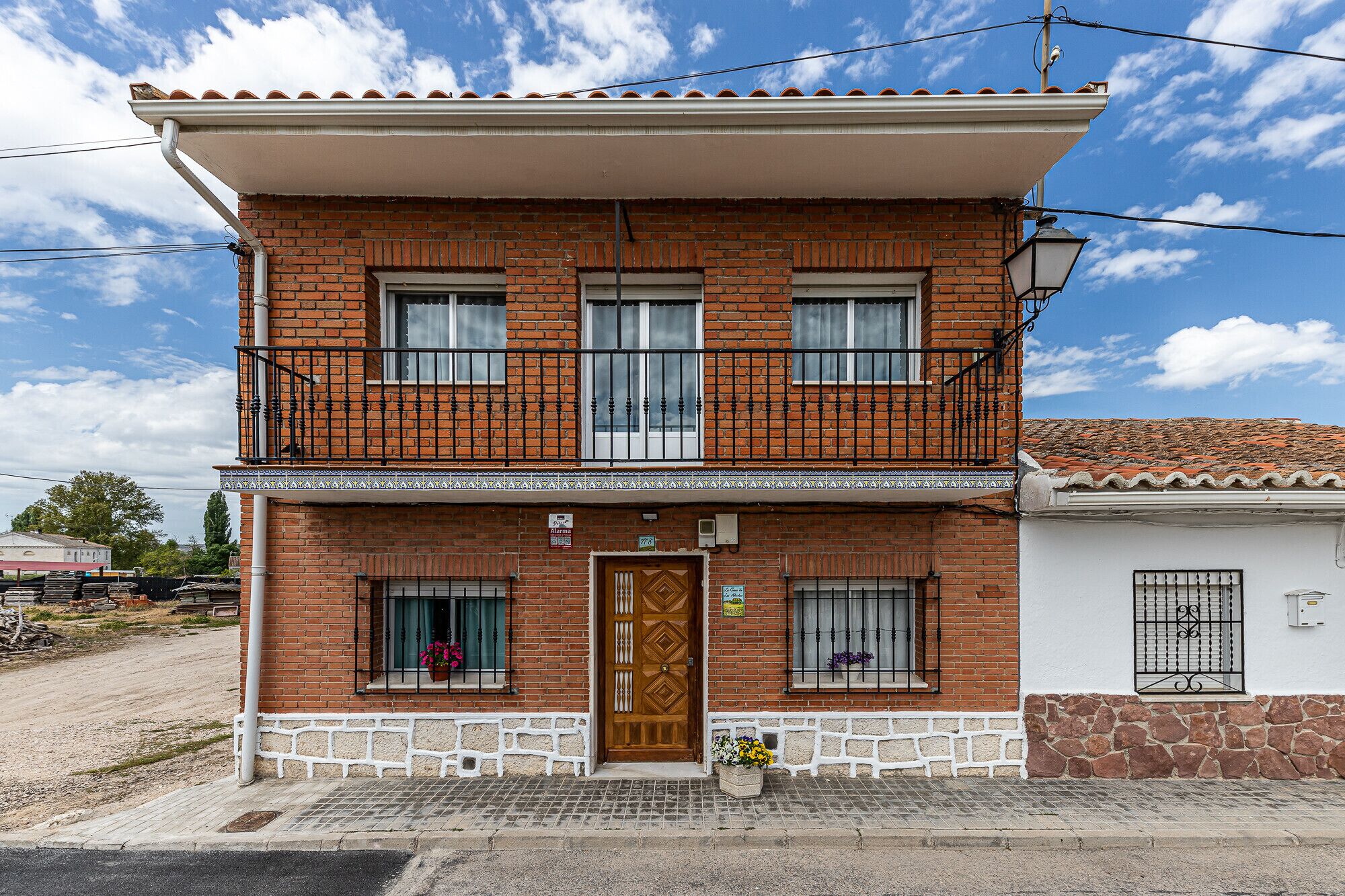 A two-story brick building with a balcony, a wooden door, and a small garden area with flowers.