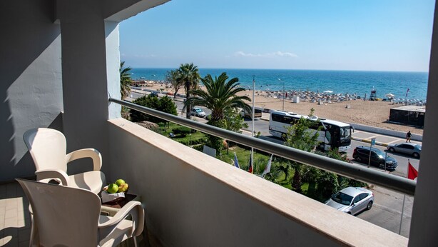 A balcony with a view of a beach, parked vehicles, and a clear sky. - FİLA OTEL AYVALIK (Ayvalik)