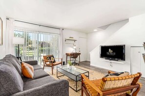 A modern living room with a sofa, chairs, a coffee table, a TV, and a view through large windows.