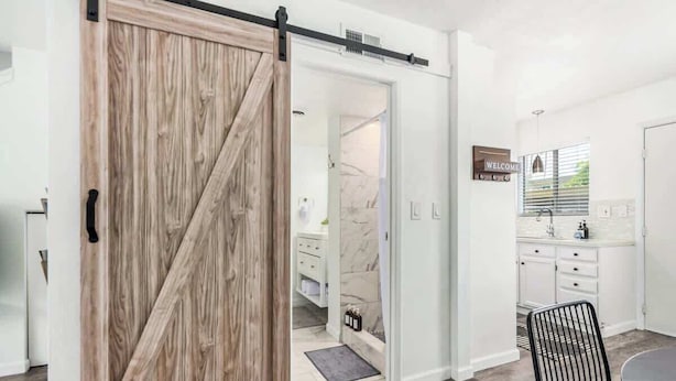 A modern kitchen with white cabinets, a black chair, and a sliding barn-style door leading to a bathroom. - Camelback 446 | Walk 2 Old Town | 4 Pools (Scottsdale)