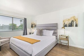 Une chambre d’hôtel moderne avec un grand lit, une table de chevet, une chaise et une fenêtre avec des stores.