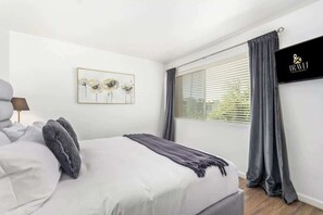 Une chambre à coucher avec un lit, un tableau, une fenêtre avec des rideaux et un téléviseur fixé au mur.
