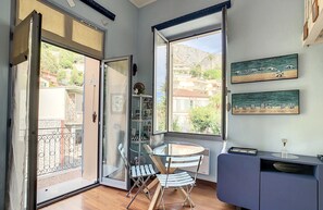 A room with a glass door leading to a balcony, a round table with a chair, a small cabinet, and a painting of a beach scene.