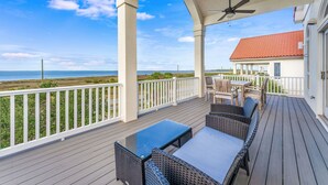 A covered outdoor deck with a view of the ocean, a white railing, and a building with a red roof. - Blue Heron - Dog Friendly, Community Pool, Across from Beach, Sunset Beach! (St. George Island)