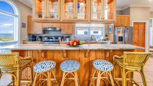 A kitchen with wooden cabinets, a central island with stools, and a view through the window. - Blue Heron - Dog Friendly, Community Pool, Across from Beach, Sunset Beach! (St. George Island)