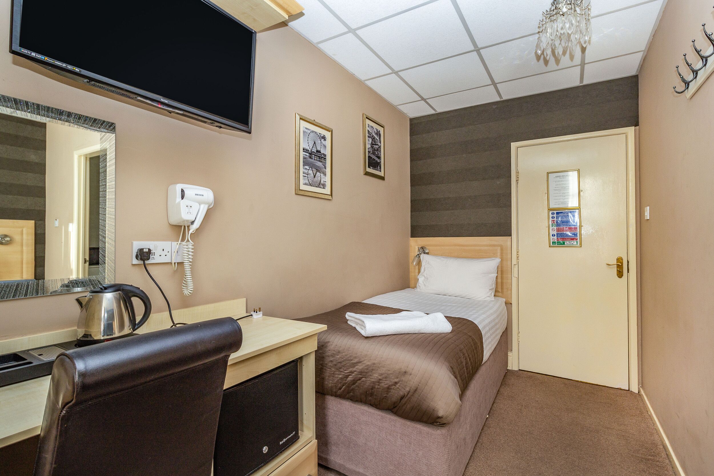 A hotel room with a bed, a desk with a kettle, a hairdryer, a television, and a door with a sign.