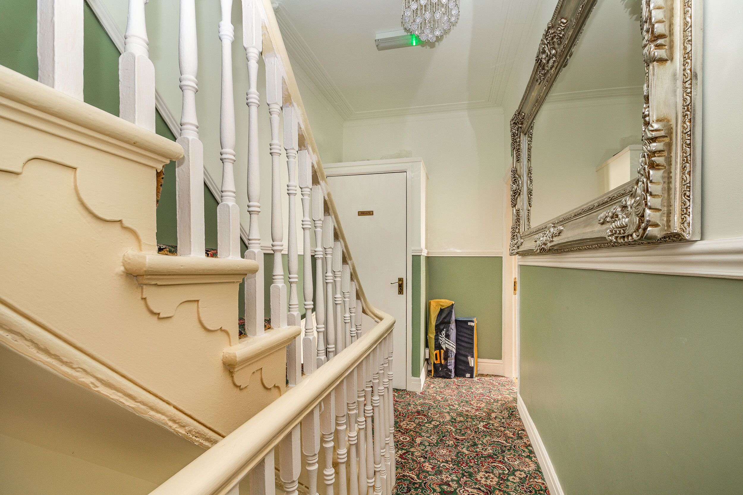 A staircase with white balusters and a patterned carpet leading to a room with a green wall and a large ornate mirror.