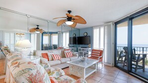 Un salon spacieux avec un grand canapé, une table basse et un téléviseur. Il y a des ventilateurs de plafond, un balcon avec des sièges d’extérieur et une vue sur l’océan.