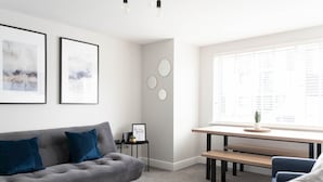 A modern living room with a grey sofa, blue cushions, a small side table with a laptop and a plant, a wooden bench, and a large window with blinds.