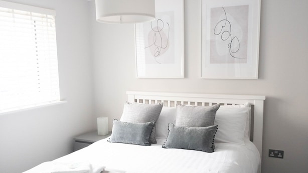 A neatly arranged bedroom with a white bed, grey pillows, and a white headboard. There are two framed artworks on the wall and a bedside table with a lamp.