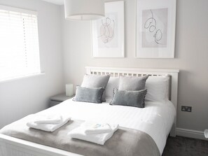 A neatly arranged bedroom with a white bed, grey pillows, and a white headboard. There are two framed artworks on the wall and a bedside table with a lamp. - Lovely, comfortable 2-bed Braehead - free parking - Pass the Keys (Renfrew)