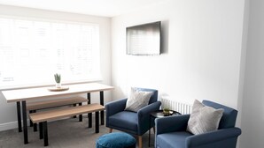 A modern living room with a white wall, a wooden table, two blue armchairs, a bench, and a small round ottoman.
