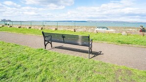 Un banco de parque frente a la orilla, con una cerca metálica y una vista distante de un horizonte urbano.