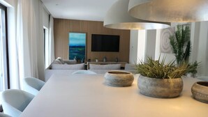 Un salon moderne avec une longue table blanche, des sièges confortables, un grand téléviseur et des pots de fleurs décoratifs.