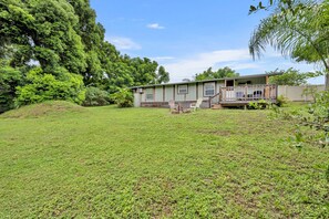 A single-story house with a front porch and a small deck, surrounded by a grassy yard and dense trees.
