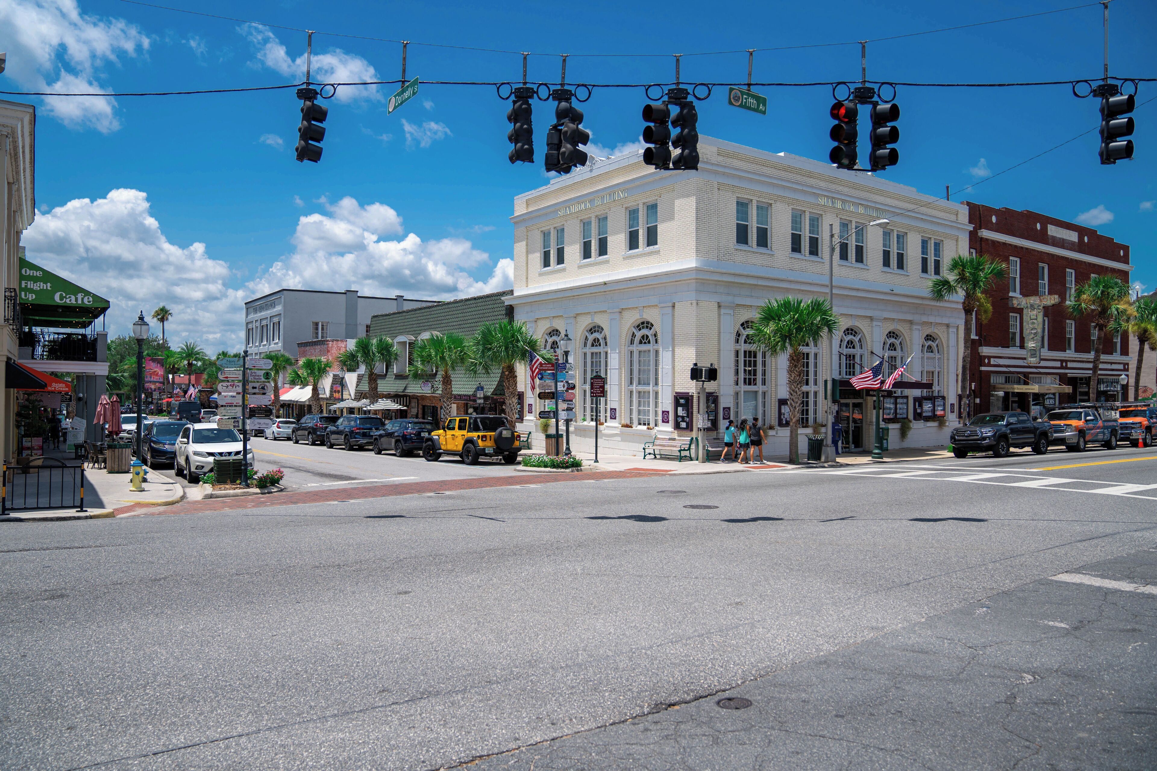 A street intersection with traffic lights, a white building with arched windows, and palm trees.
