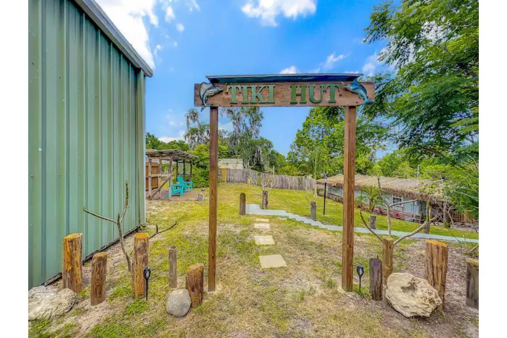 Une enseigne en bois portant l’inscription « TIKI HUT », placée dans un décor extérieur rustique, avec un bâtiment vert, des poteaux en bois et une structure à toit de chaume.